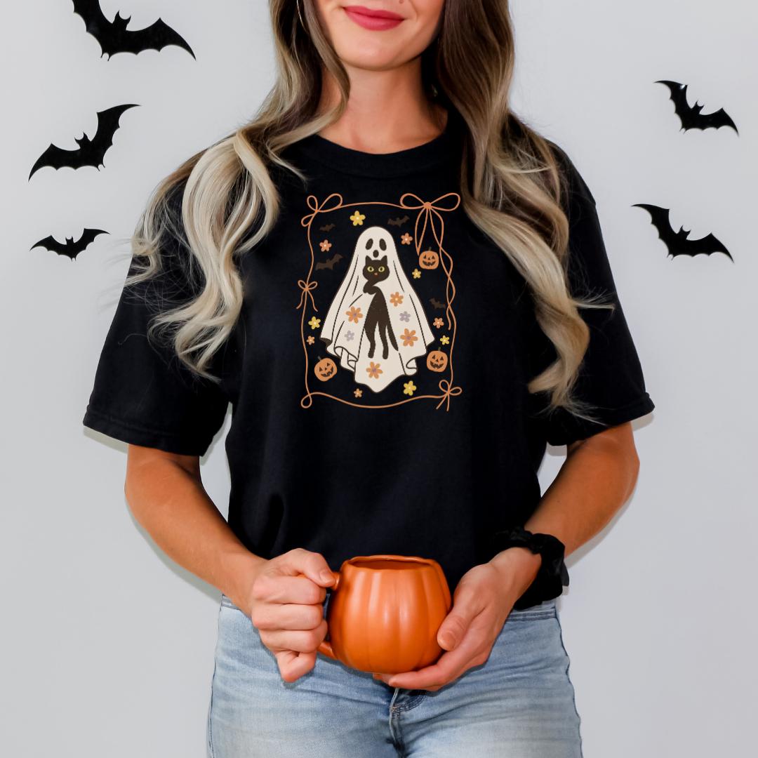 A woman wearing a black t-shirt with a Halloween-themed design, including a ghost, bats, and a pumpkin, is holding a small orange mug.