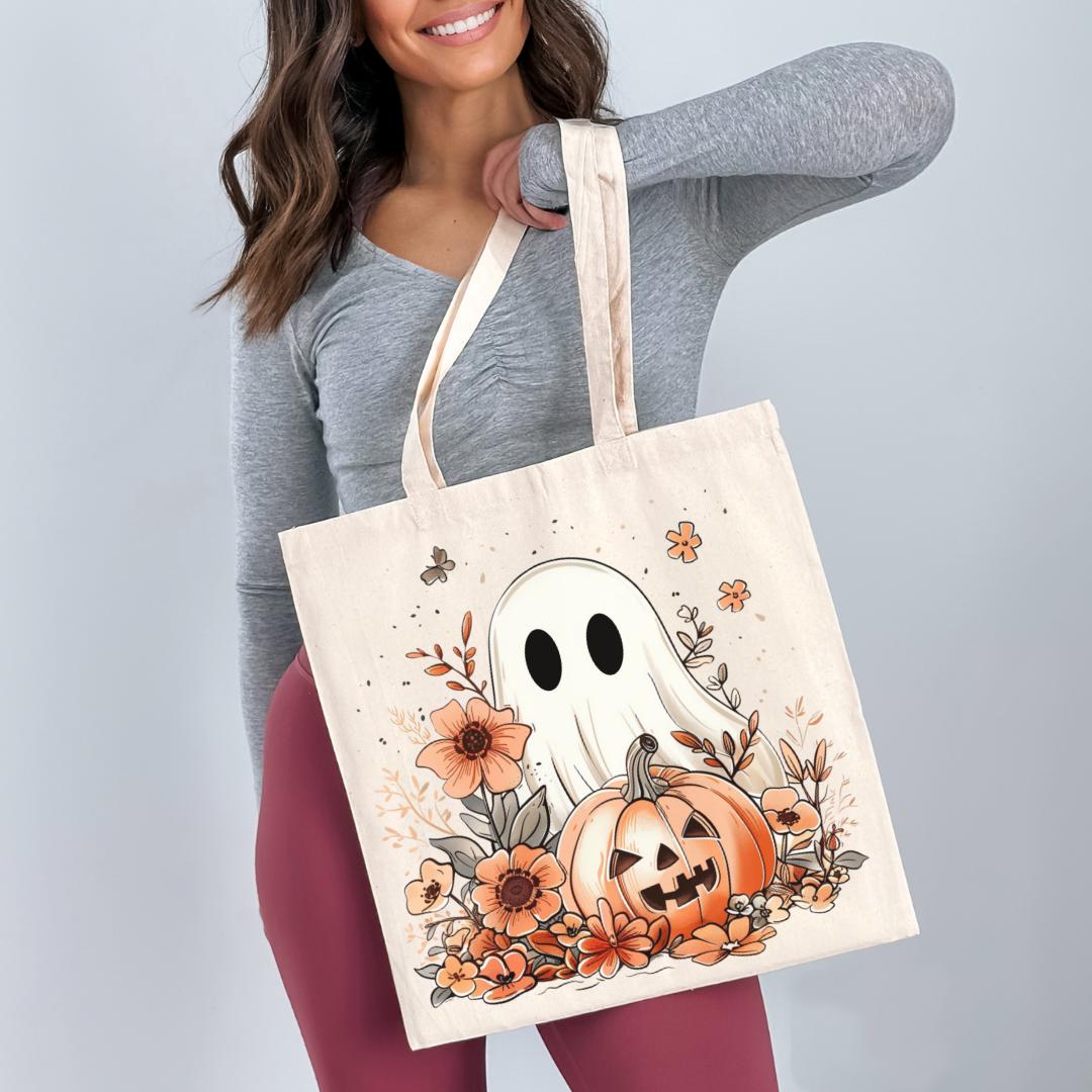 A woman is holding a white tote bag with a Halloween-themed design featuring a ghost, pumpkin, and flowers.