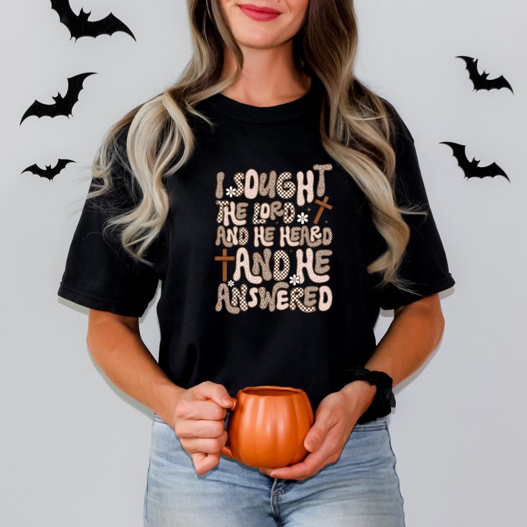 A woman with long blonde hair wearing a black t-shirt with a Halloween-themed design holds a pumpkin-shaped mug.