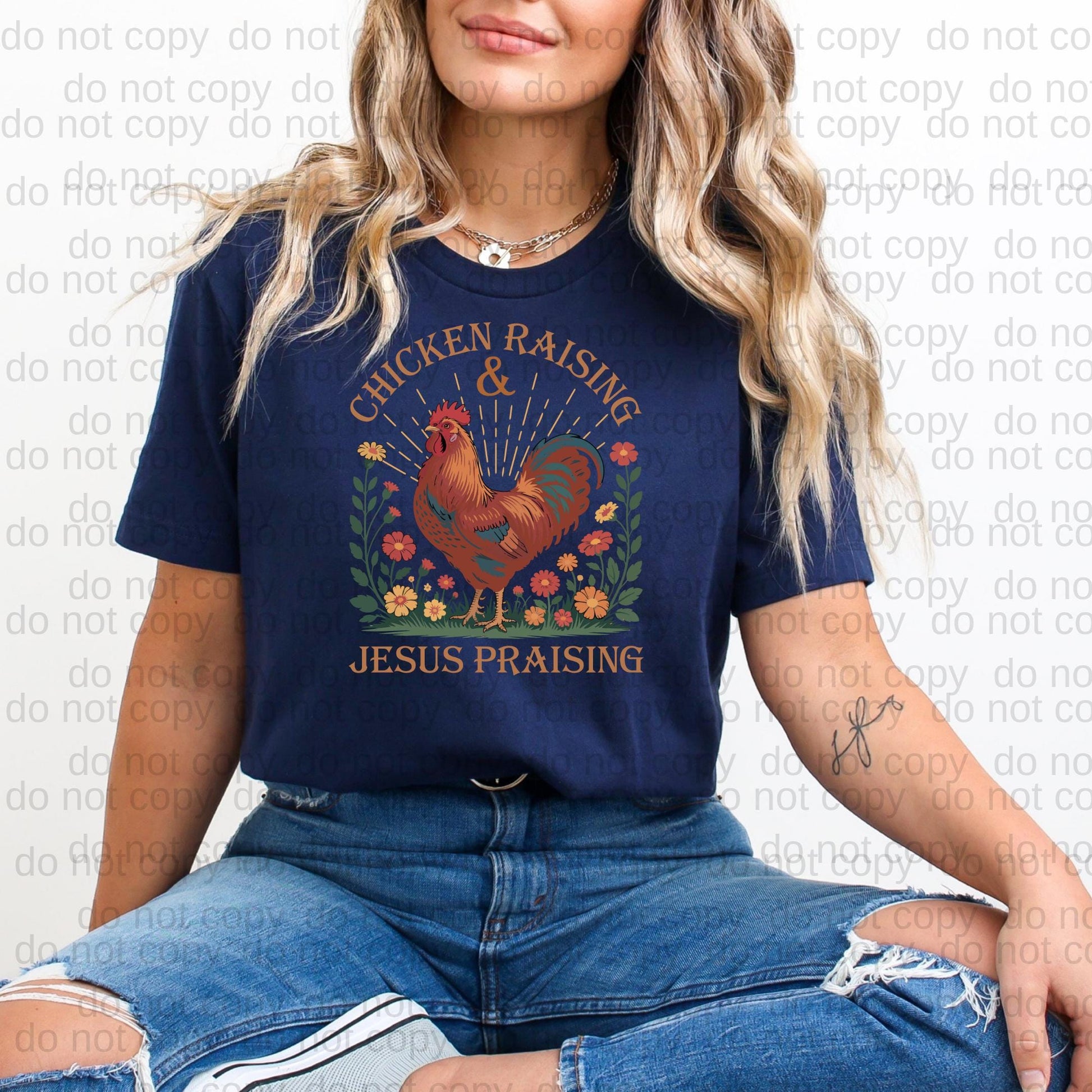 A woman wearing a navy blue t-shirt with a graphic design of a rooster and flowers, sitting on the ground with her legs crossed.