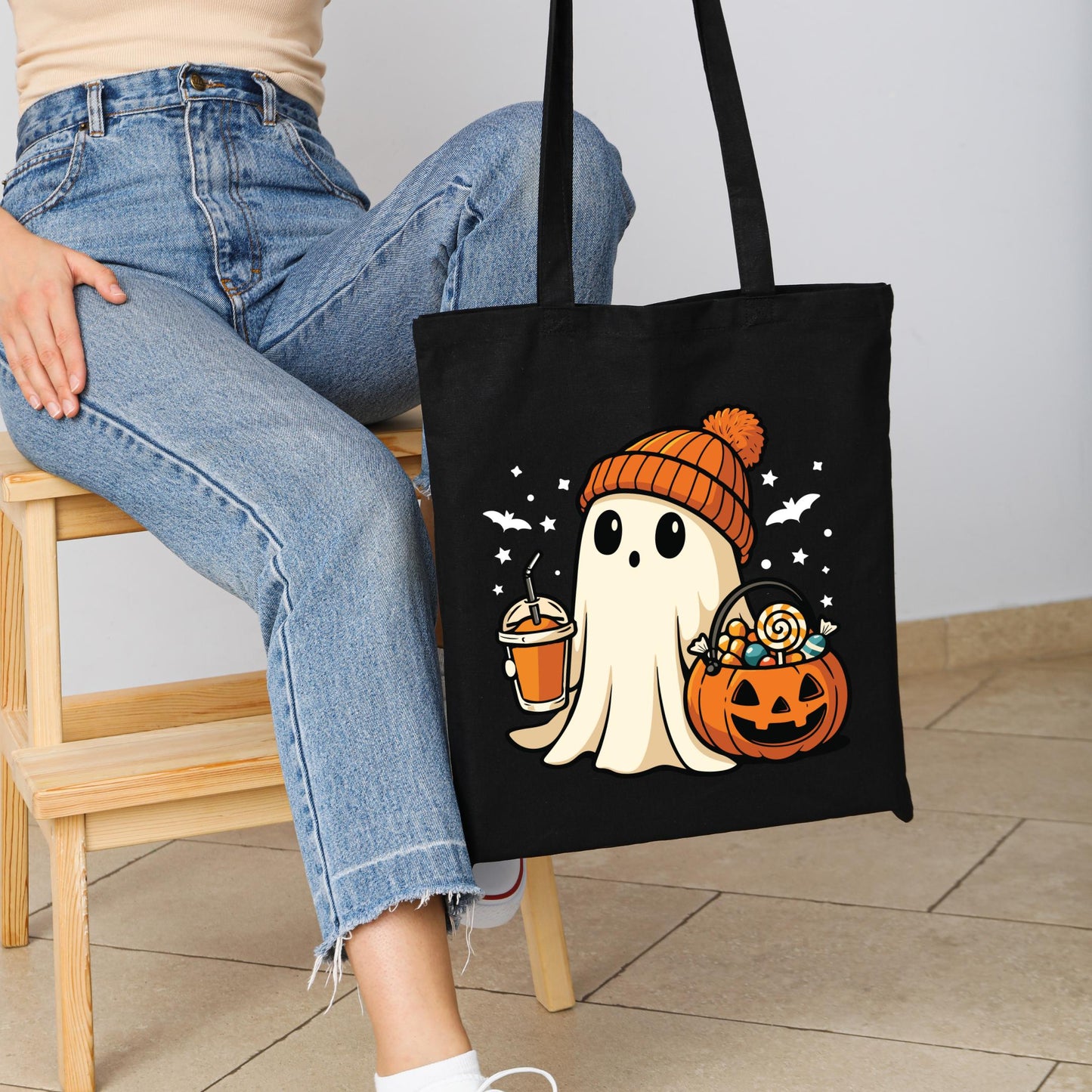 A person is sitting on a wooden chair, holding a black tote bag featuring a cartoon ghost character wearing a hat and holding a pumpkin.