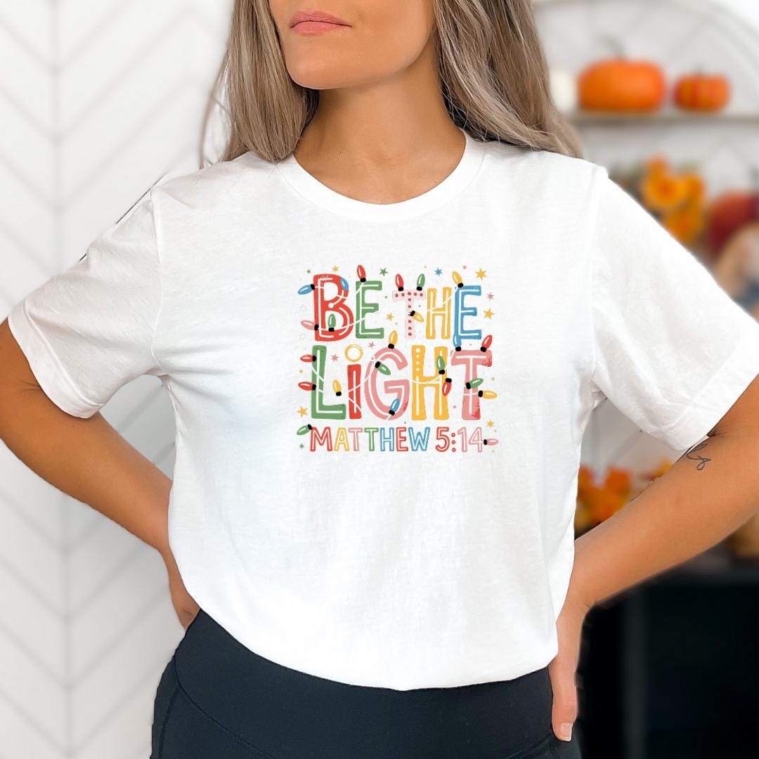 A woman wearing a white t-shirt with a colorful graphic design featuring the text "Be the light" and "MATTHEW 5:14".