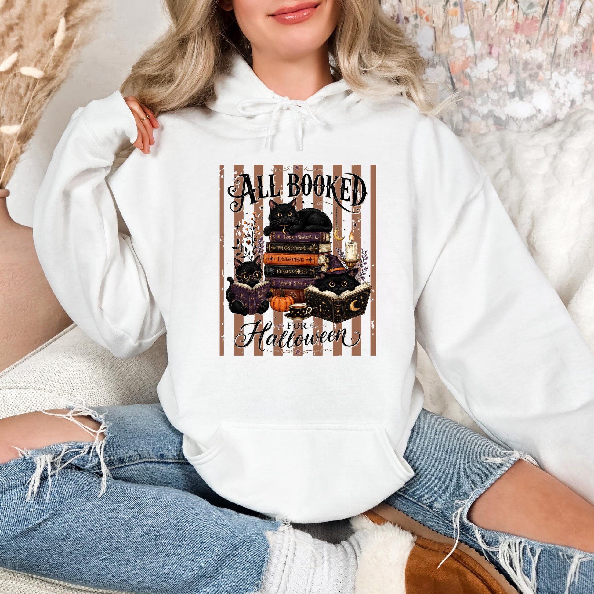 A woman wearing a white hoodie with a graphic design of a stack of books and cats, along with the text "All Booked for Halloween".