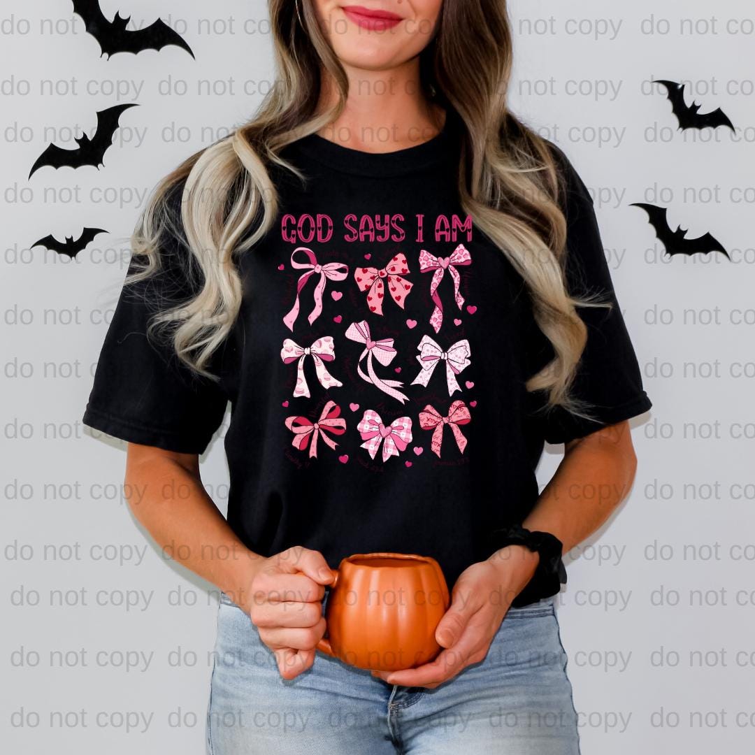 A woman wearing a black t-shirt with a graphic design of pink bows and the text "GOD SAYS I AM" is holding a pumpkin.