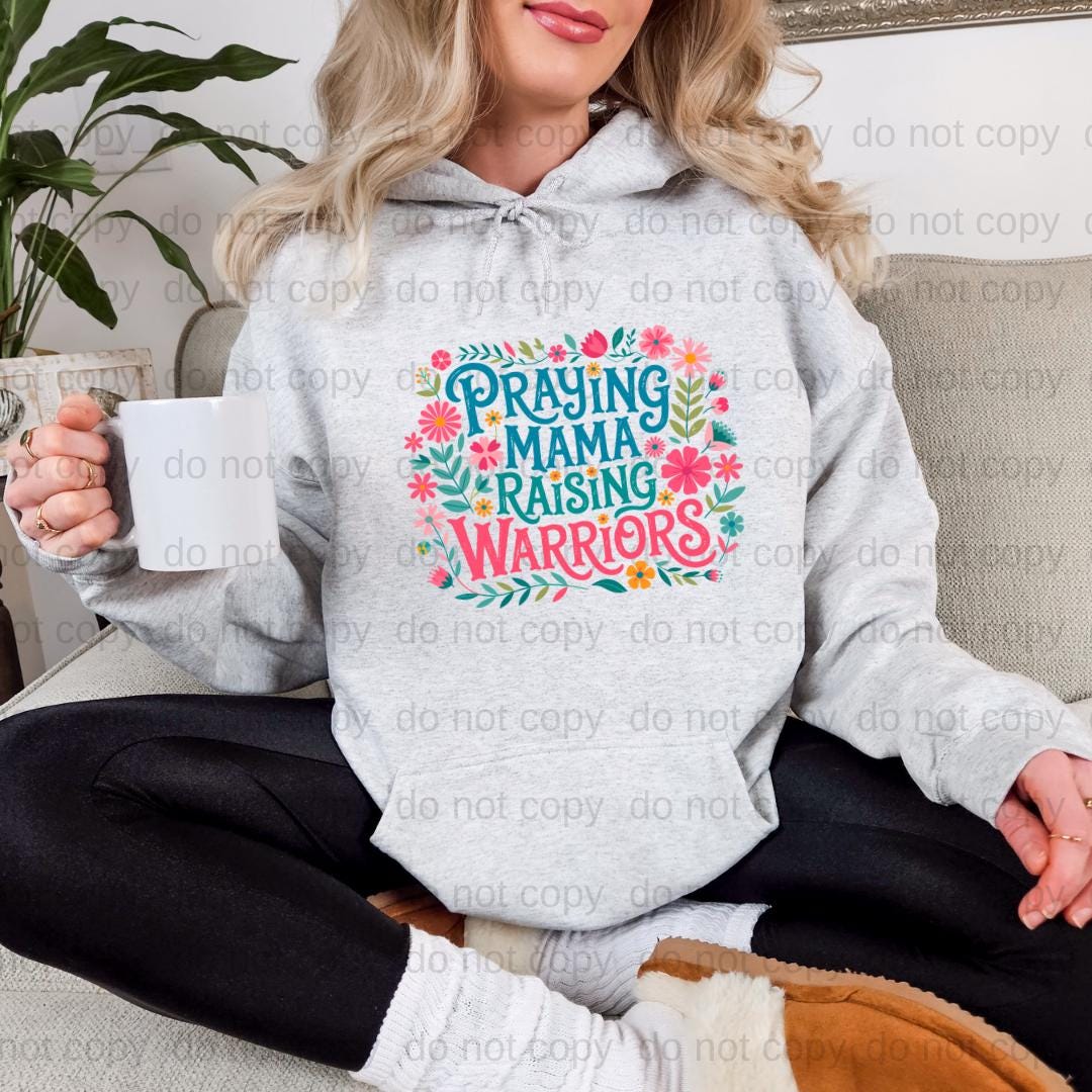 A woman sitting on a couch wearing a gray hoodie with a colorful graphic design that says "Praying Mama Raising Warriors".