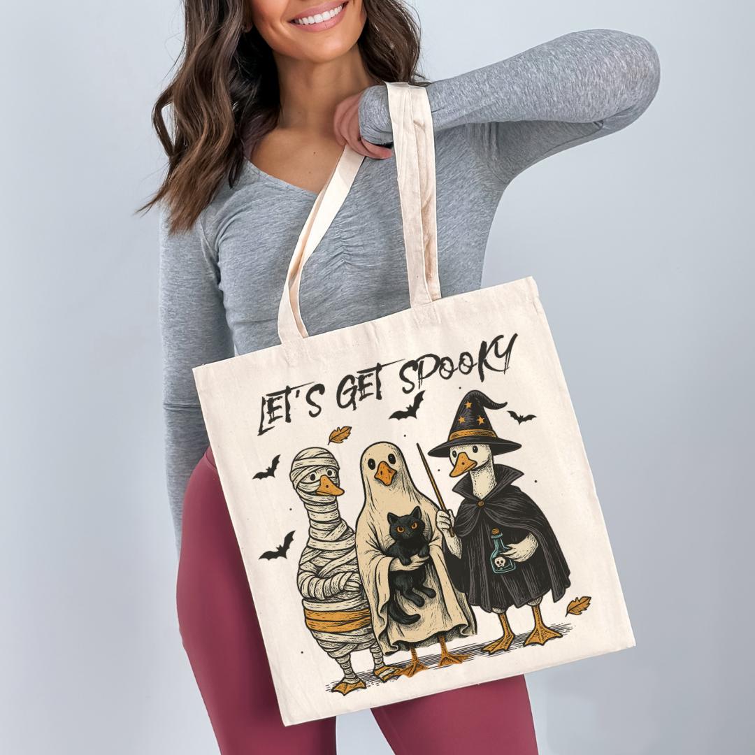 A woman is holding a tote bag with a Halloween-themed illustration of three cartoon ducks dressed in costumes, including a mummy and a witch, with the text "Let's get spooky" printed on it.