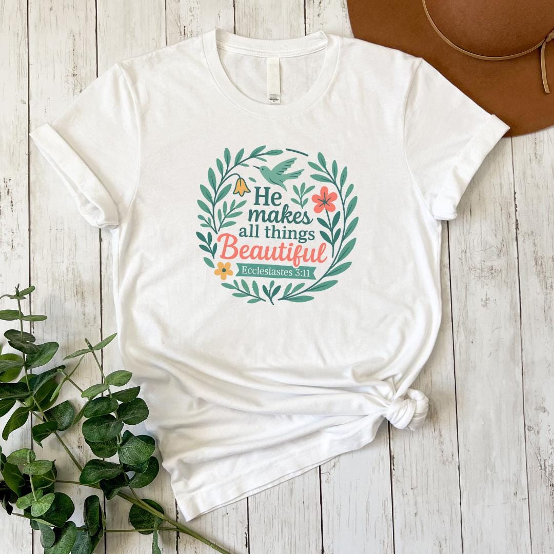 A white t-shirt with a colorful graphic design and text, along with a brown hat and some greenery, are arranged on a wooden surface.
