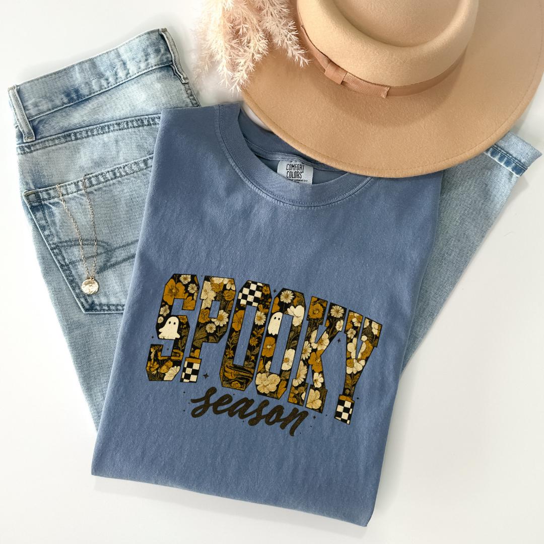 A casual outfit consisting of a blue t-shirt with a floral print, a pair of jeans, and a beige hat is laid out on a white surface.
