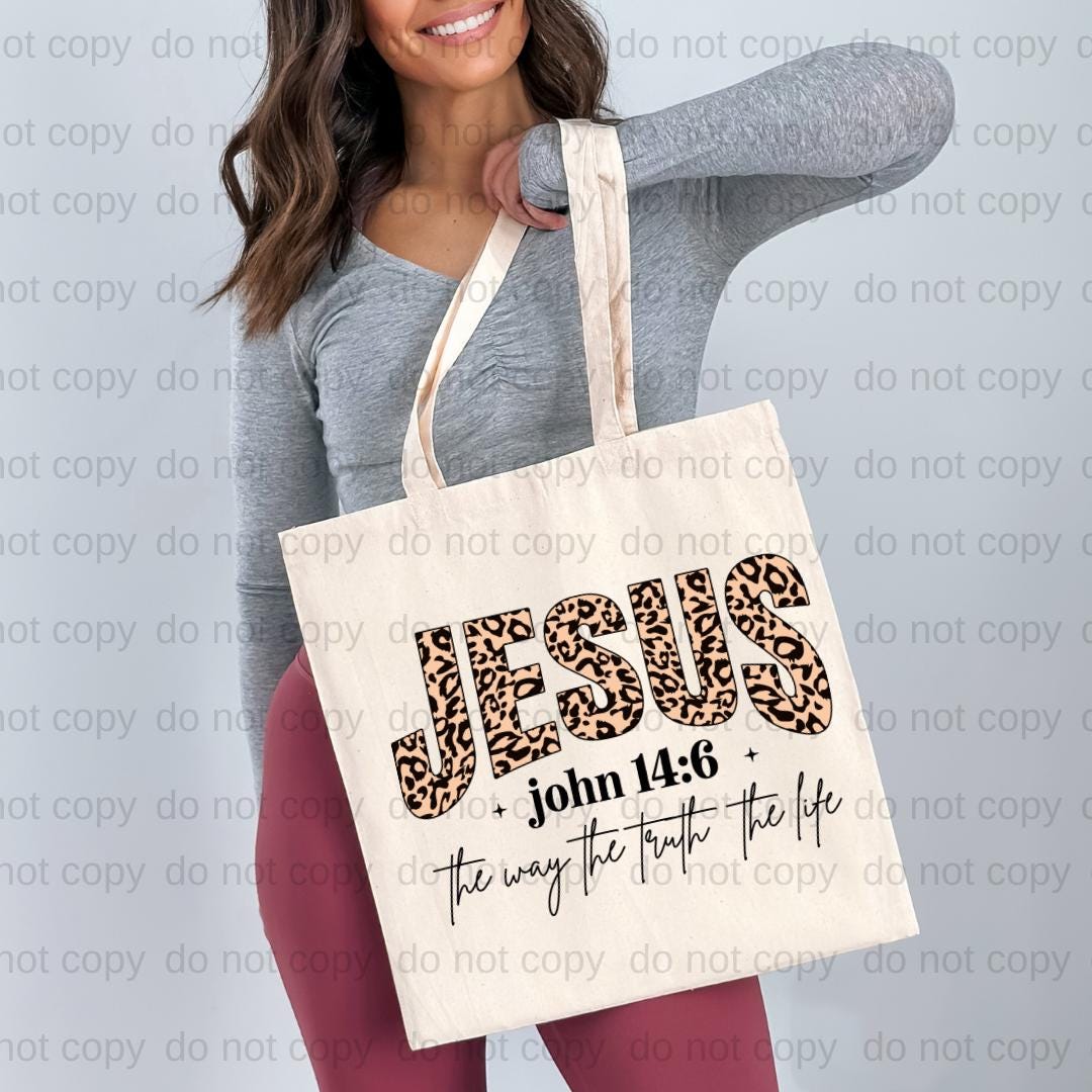 A woman is holding a white tote bag with the text "JESUS" and "John 14:6" printed on it.