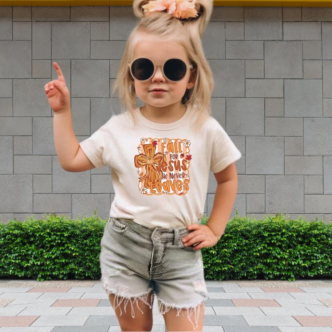 A young girl wearing a white t-shirt with a graphic design, sunglasses, and denim shorts is standing in front of a brick wall.