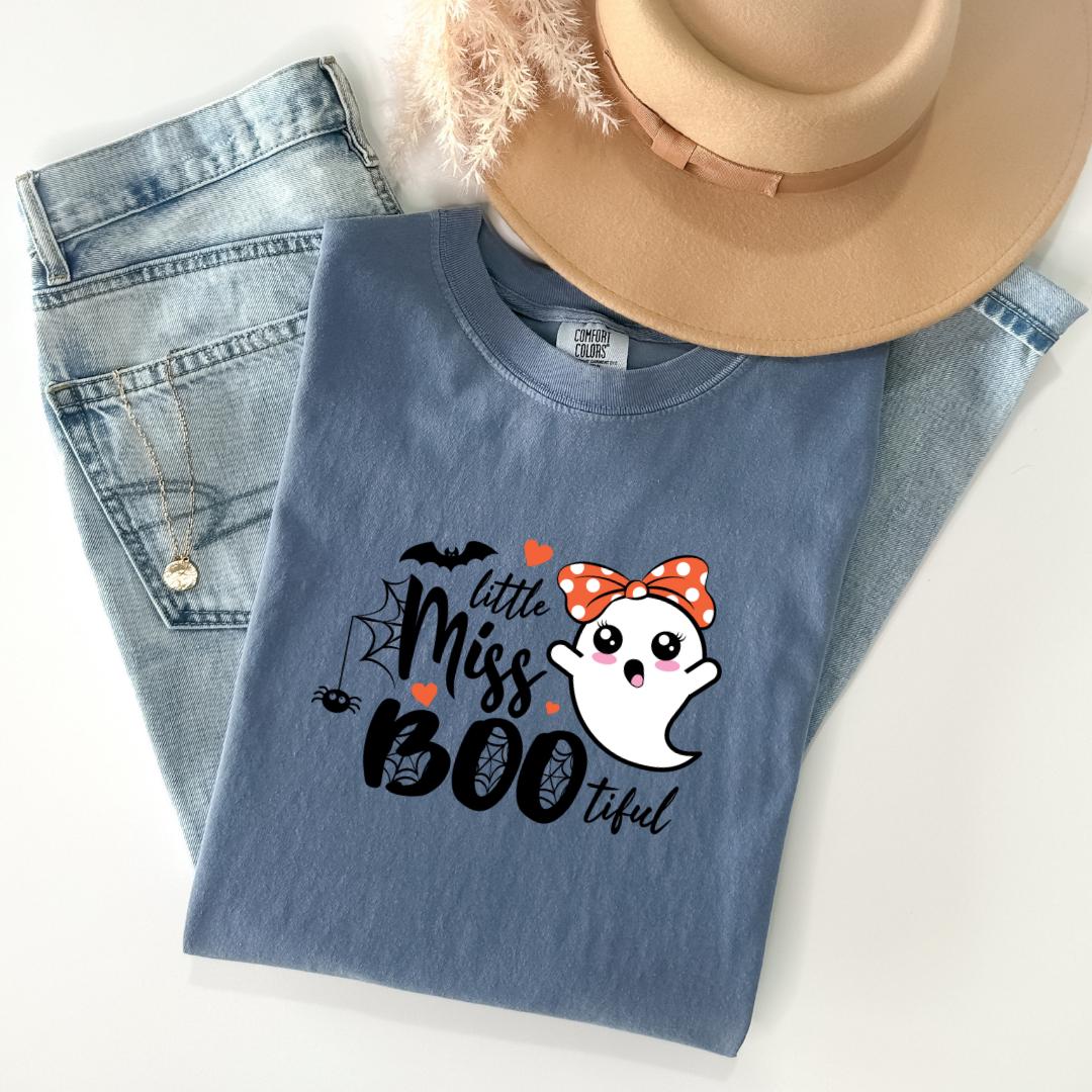 A blue t-shirt with a graphic design of a ghost wearing a red bow and the text "Little Miss BOO tful" printed on it, along with a beige hat and a pair of jeans.
