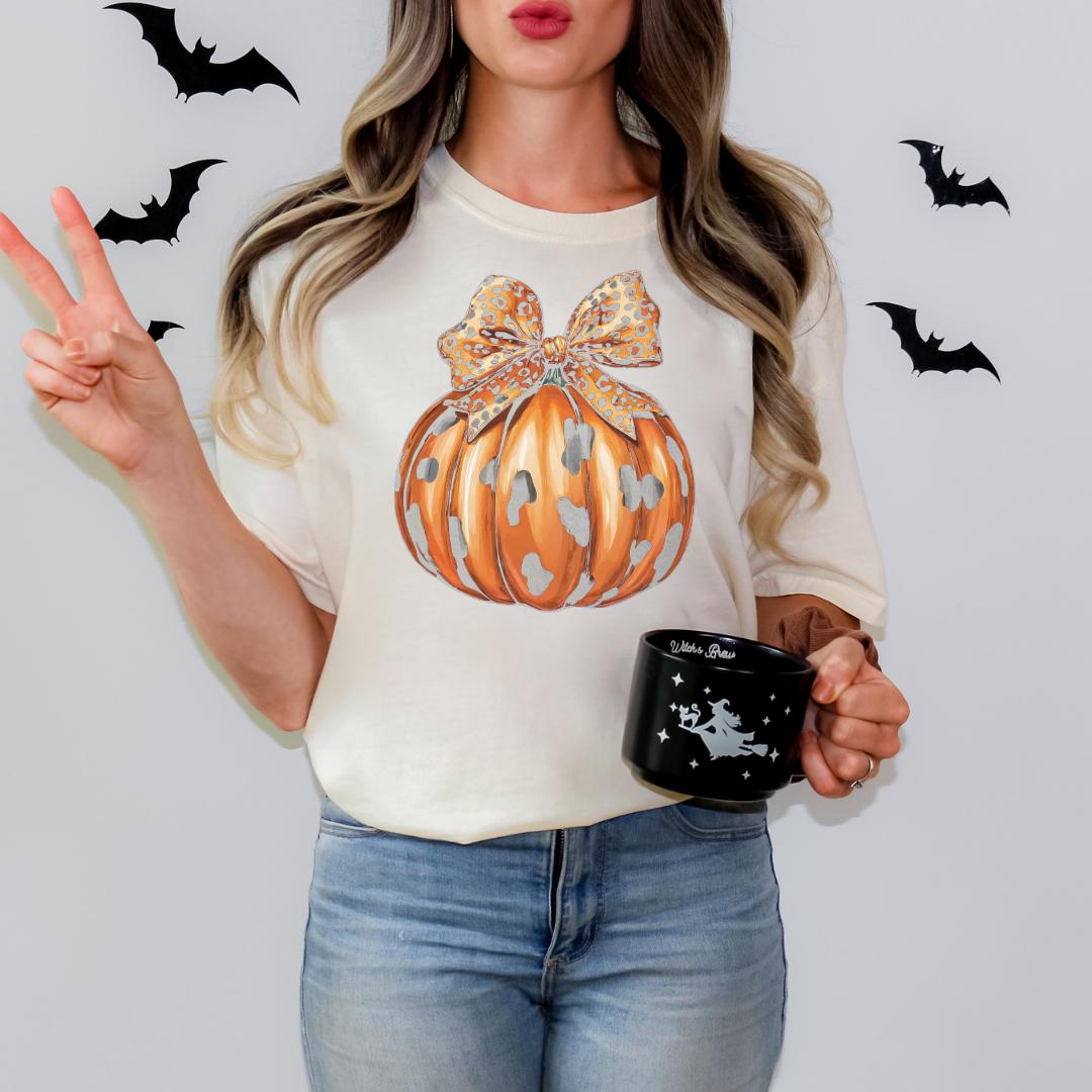 A woman is standing against a white background, wearing a white t-shirt with a pumpkin graphic and holding a black mug with a star design.
