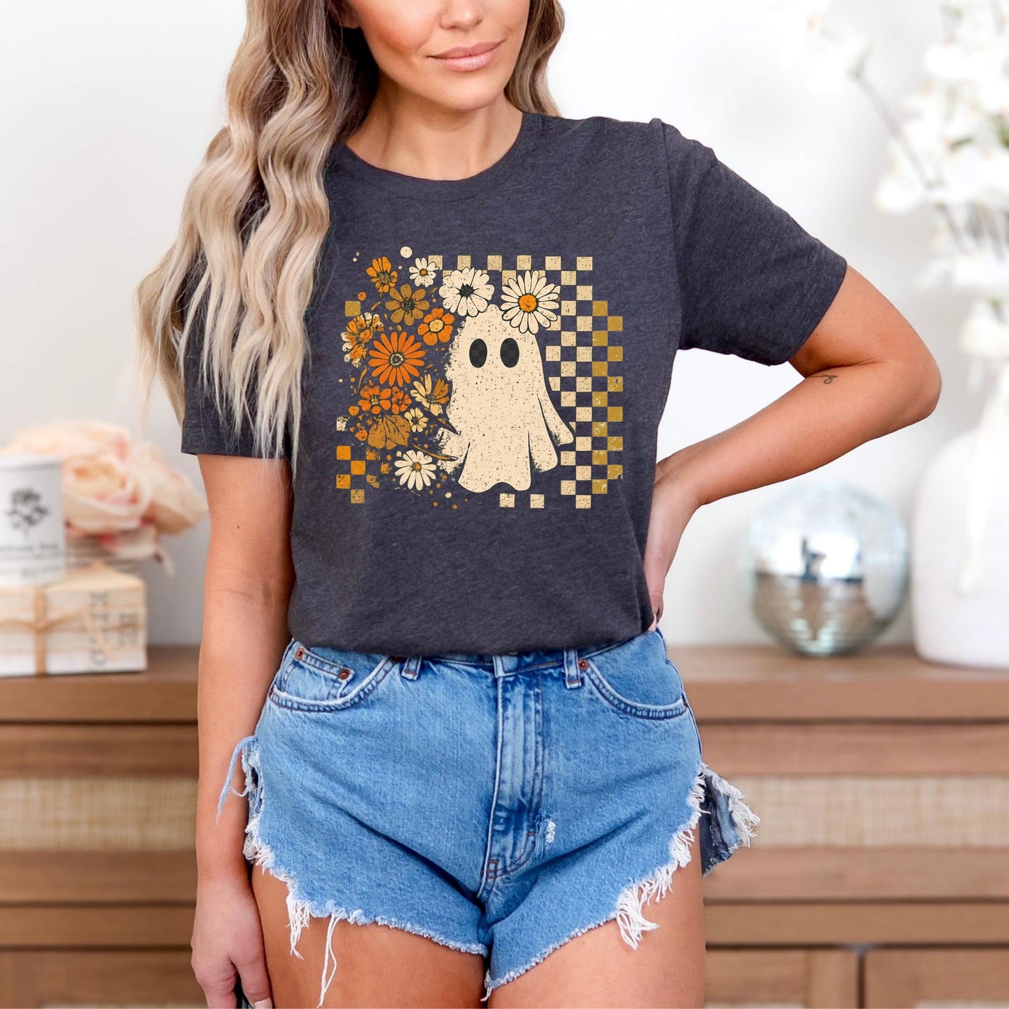 A woman wearing a black t-shirt with a graphic of a ghost surrounded by flowers and a checkered pattern, paired with denim shorts.