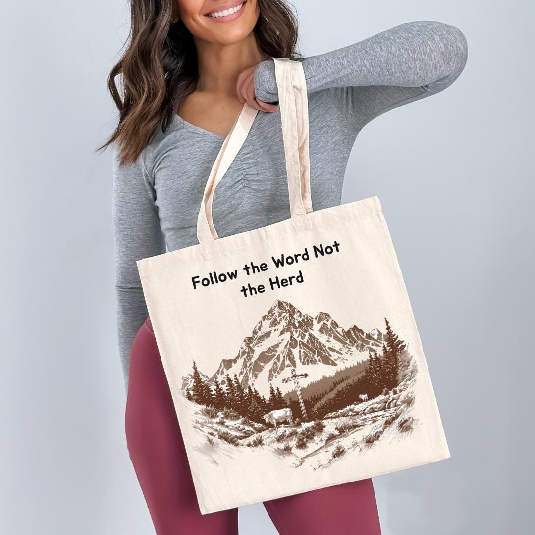 A woman holding a white tote bag with a printed design of a mountain landscape and the text "Follow the Word Not the Herd".