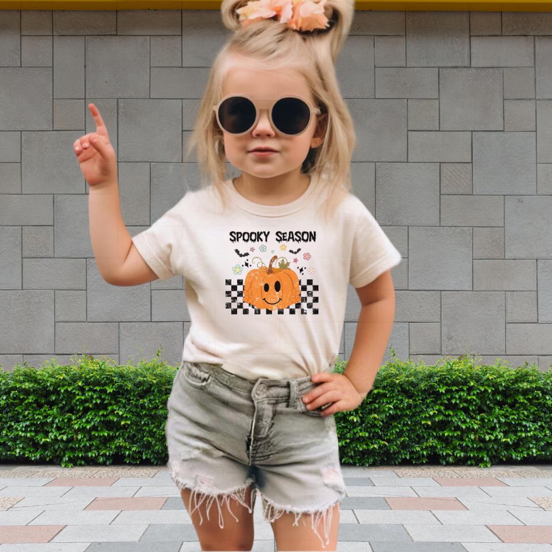 A young girl wearing a white t-shirt with a pumpkin graphic and the text "SPOKEY SEASON" is standing in front of a brick wall.