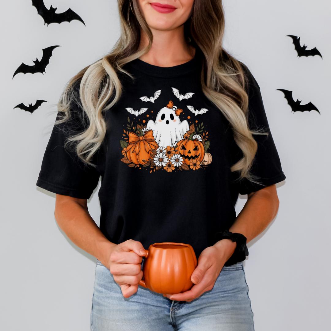 A woman wearing a black t-shirt with a Halloween-themed graphic of a ghost, pumpkin, and bats, holding a small orange pumpkin-shaped mug.