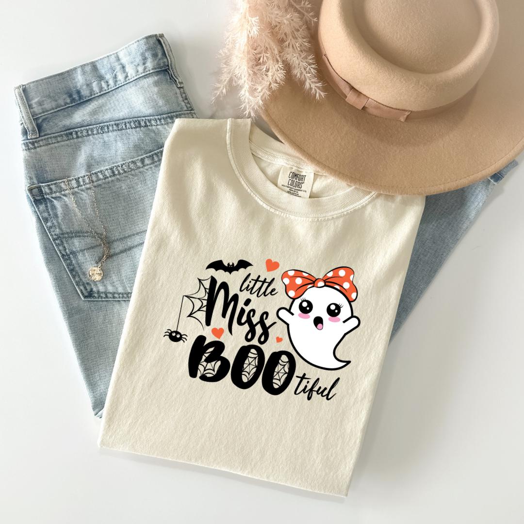 A white t-shirt with a graphic design featuring a ghost character and the text "Little Miss Boo" printed on it, along with a bow and a spider web. The t-shirt is placed on top of a pair of jeans and a beige hat.