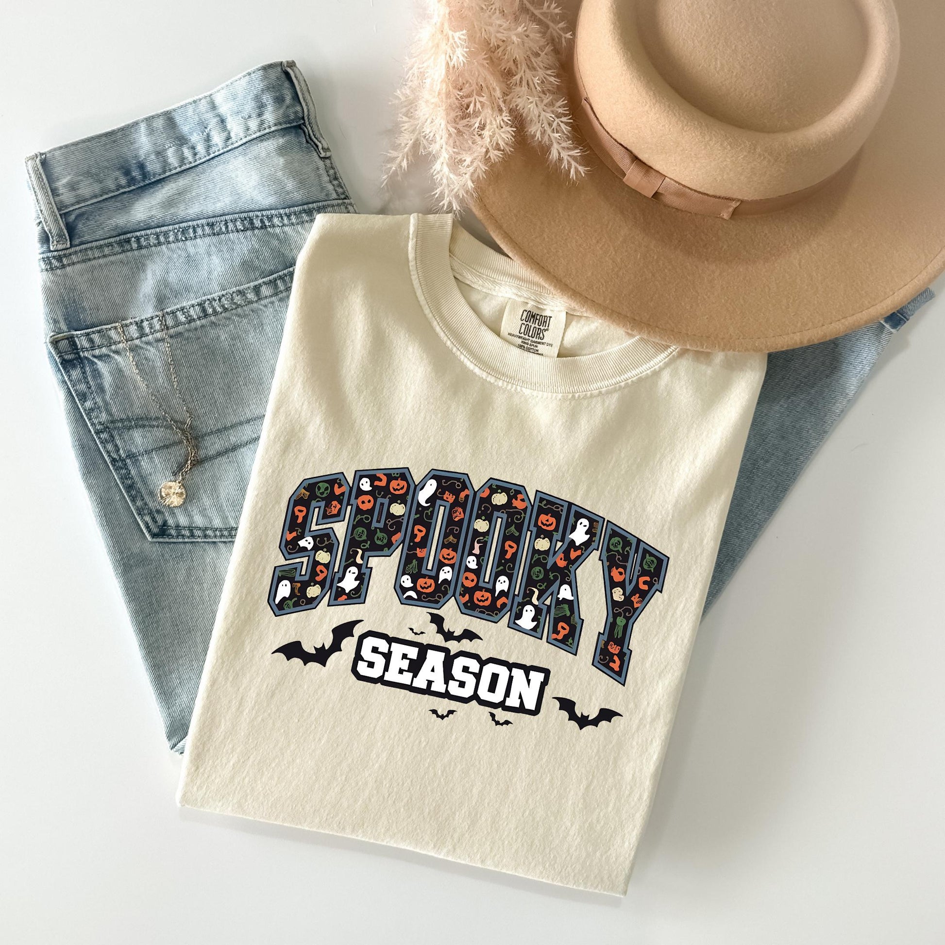 A white t-shirt with a Halloween-themed design, a pair of jeans, and a beige hat are arranged on a white surface.