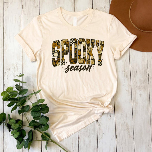 A white t-shirt with a floral print design and the text "SPOKY season" printed on it, along with a brown hat placed next to it on a wooden surface.
