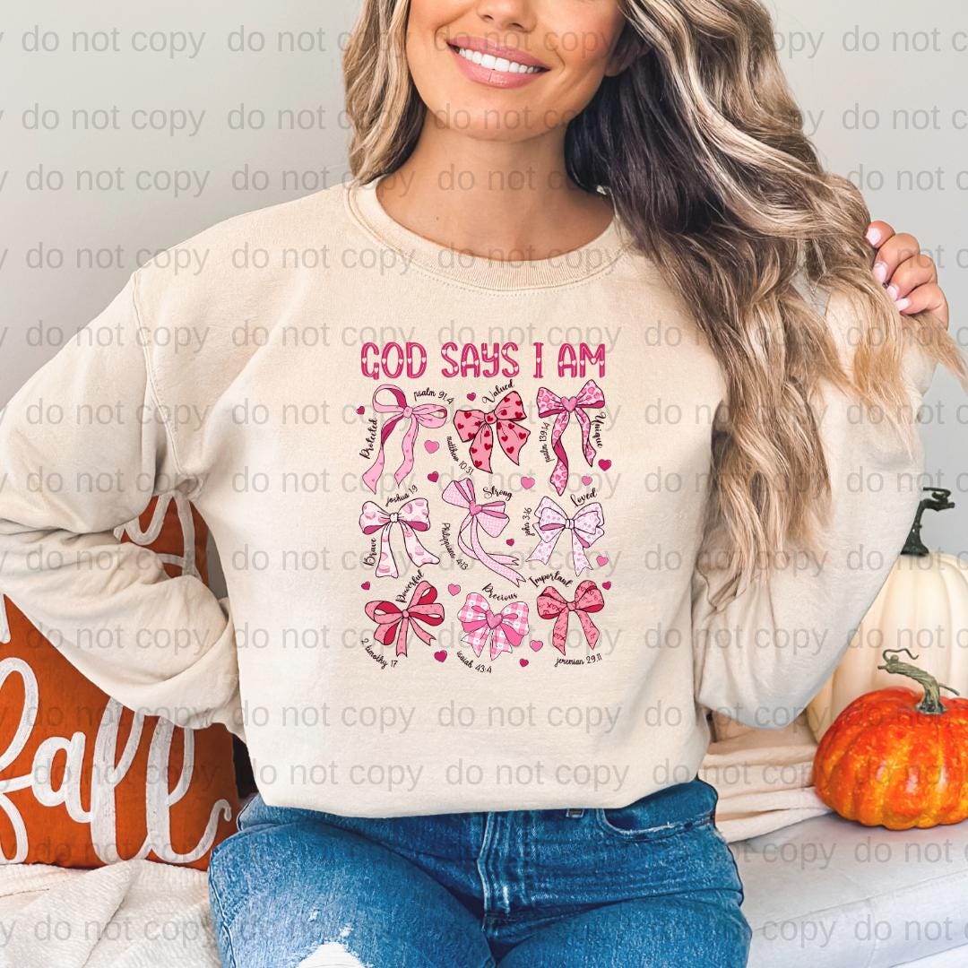 A woman wearing a white sweatshirt with a graphic design of pink bows and the text "God says I am" printed on it.