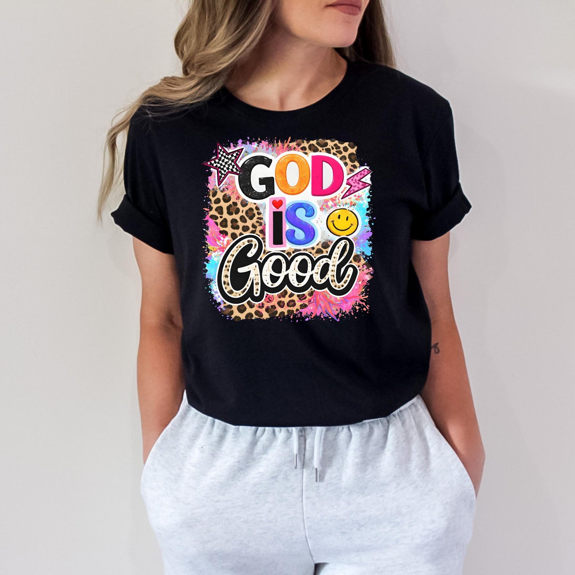 A woman wearing a black t-shirt with a colorful graphic design that says "GOOD IS GOOD" and includes a smiley face and a cheetah print.