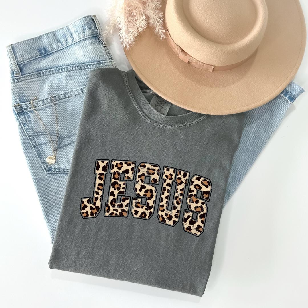 A gray t-shirt with the word "JESUS" printed on it, along with a beige hat and a pair of blue jeans, all laid out on a white surface.