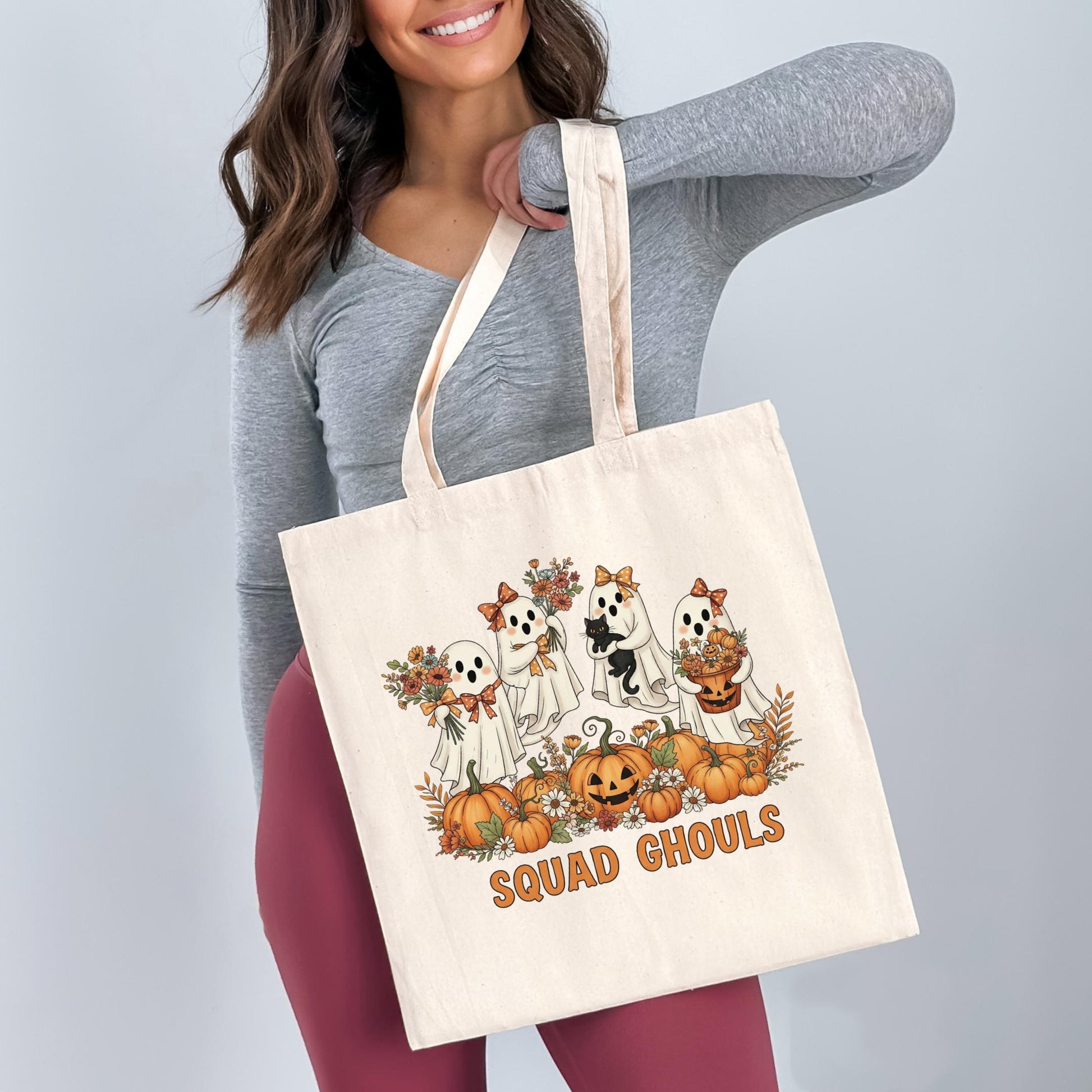 A woman is holding a white tote bag with a Halloween-themed design featuring ghostly figures and pumpkins.