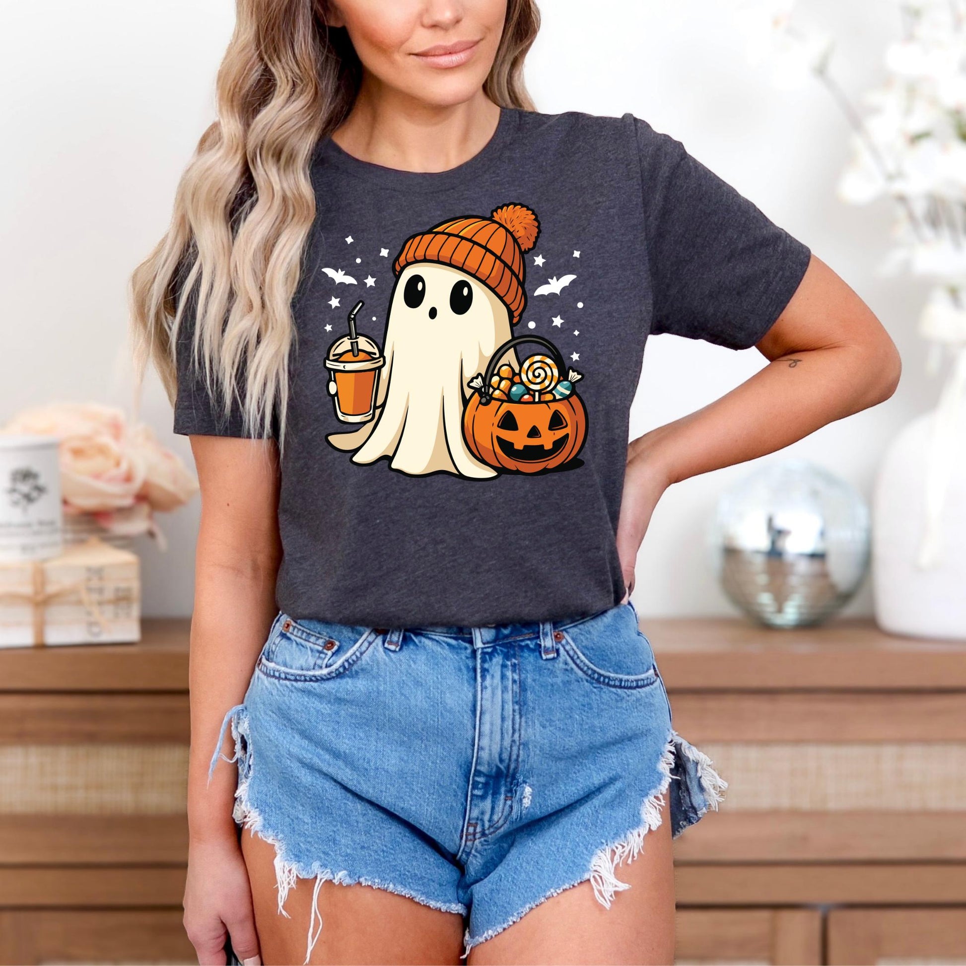 A woman wearing a black t-shirt with a graphic of a ghost holding a pumpkin and a cup, standing in front of a wooden dresser.