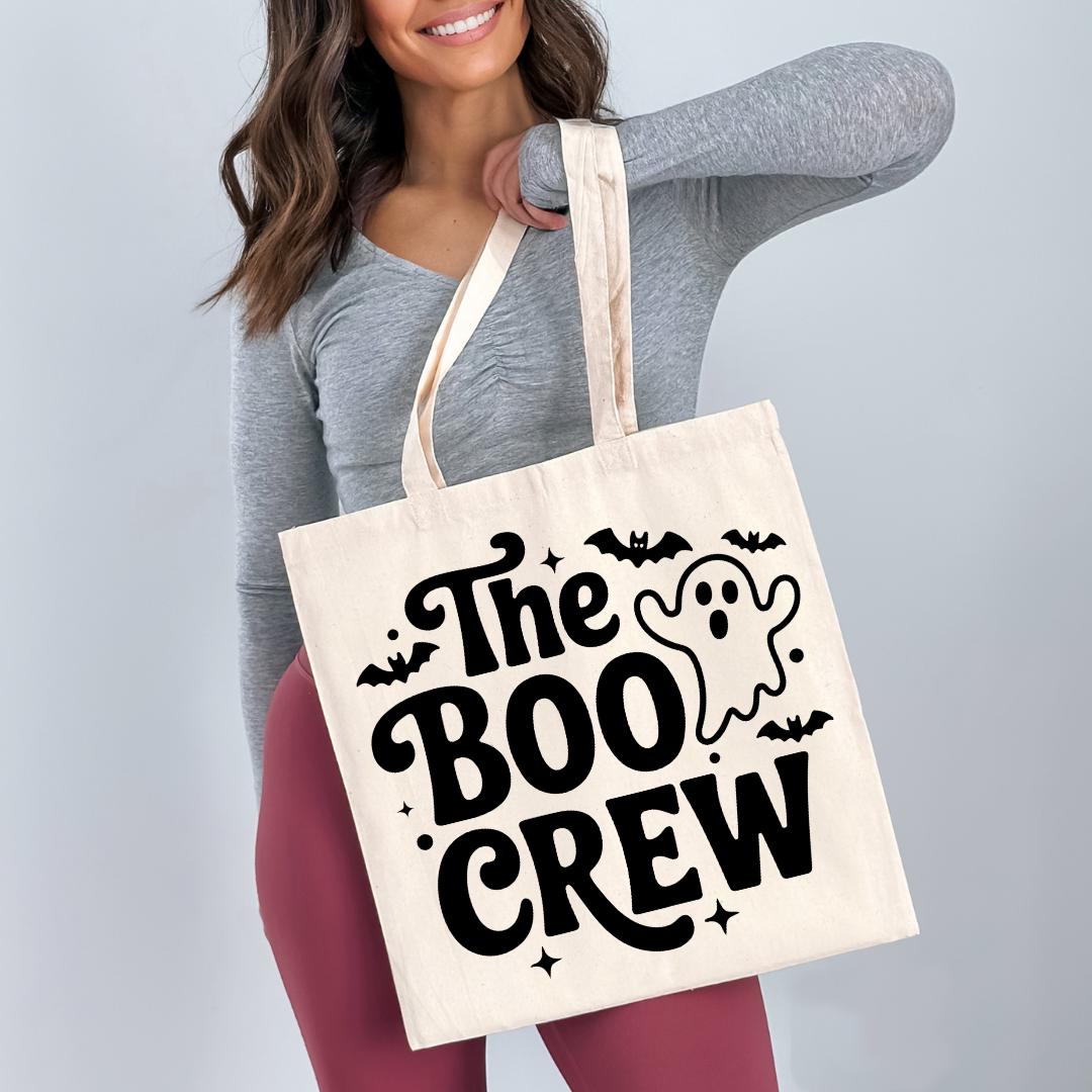 A woman is holding a white tote bag with the text "The Boo Crew" printed on it, which appears to be a Halloween-themed design.