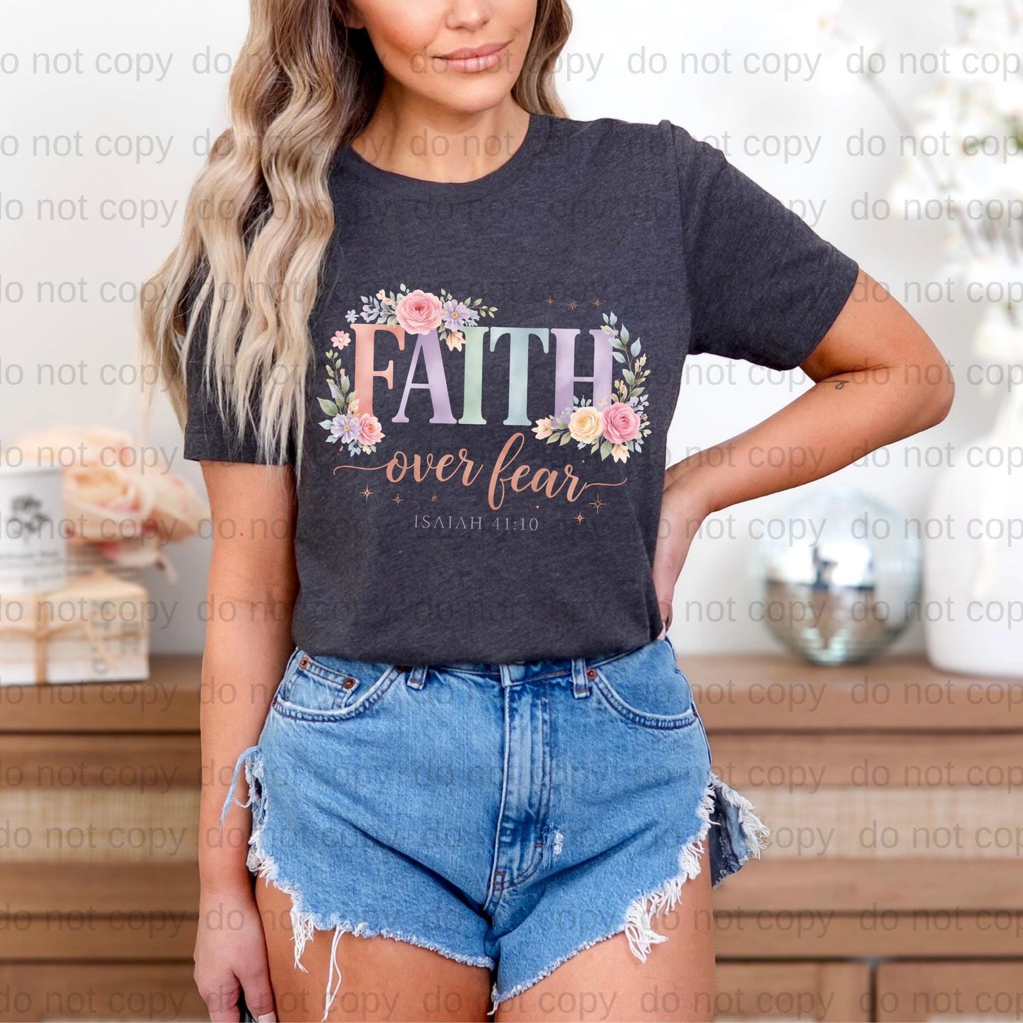 A woman wearing a gray t-shirt with a floral design and denim shorts stands in front of a wooden dresser.