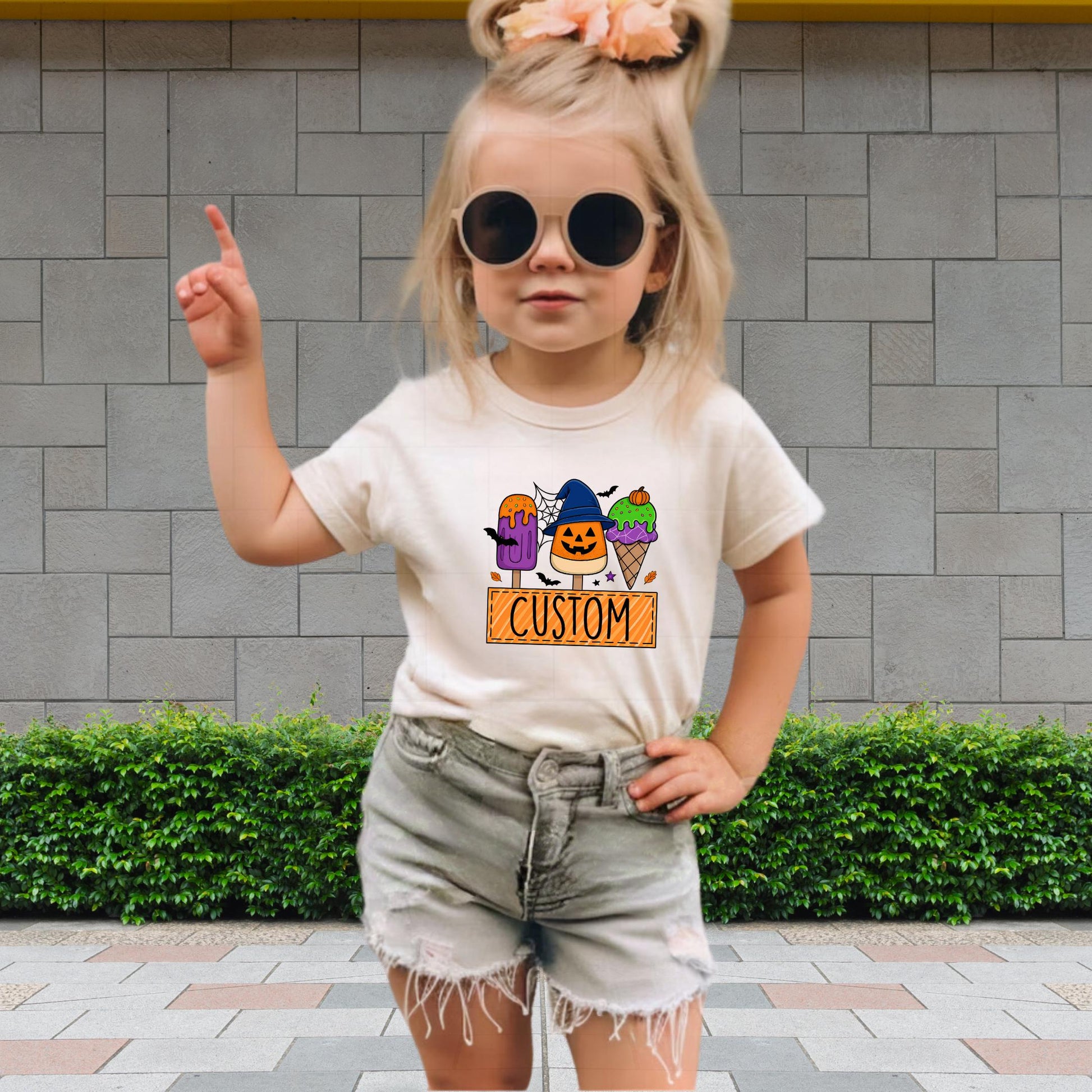 A young girl wearing a white t-shirt with a graphic design and sunglasses is standing in front of a brick wall.
