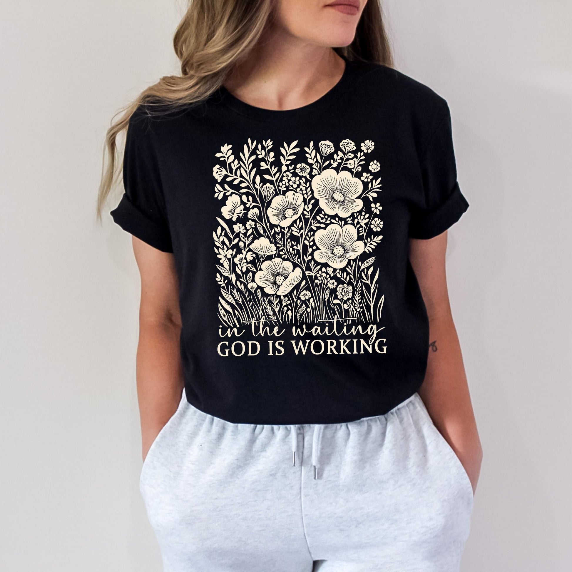 A woman wearing a black t-shirt with a floral design and the text "In the waiting, God is working" printed on it.