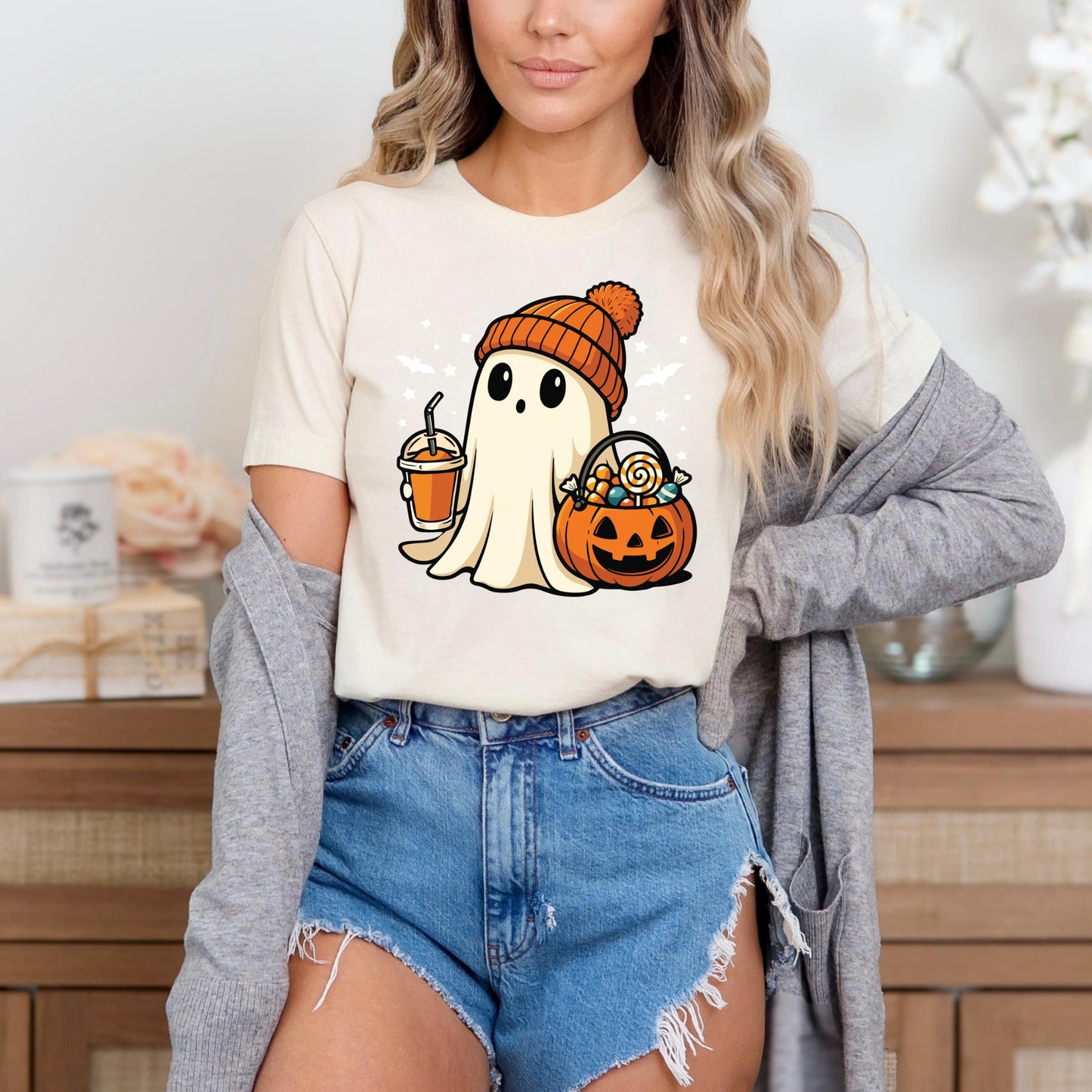 A woman wearing a white t-shirt with a graphic of a ghost holding a pumpkin and a drink, paired with denim shorts and a gray cardigan.