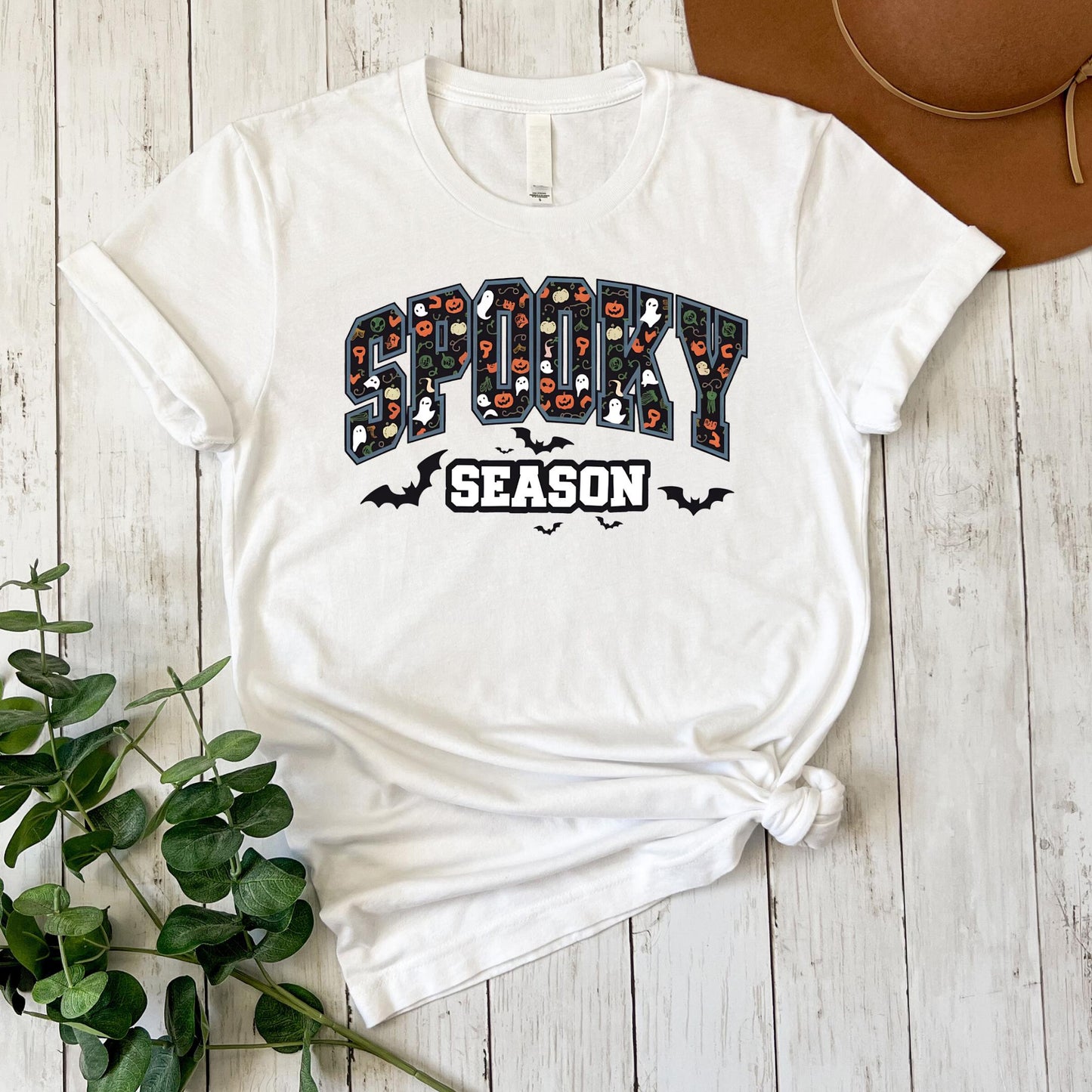 A white t-shirt with a Halloween-themed graphic design, including the text "SPOOKY SEASON" and various spooky symbols, is displayed on a wooden surface alongside a brown hat.