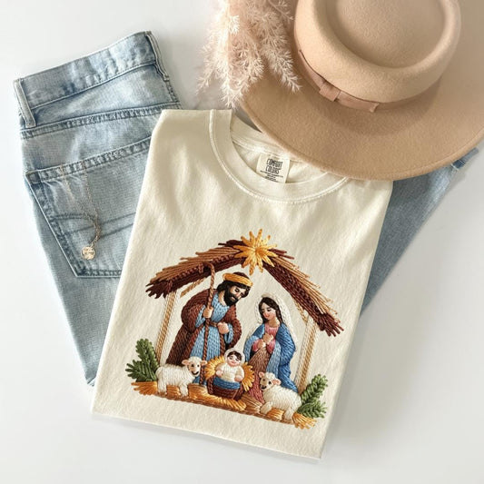 A white t-shirt with a nativity scene design, a pair of blue jeans, and a beige hat are arranged on a white surface.
