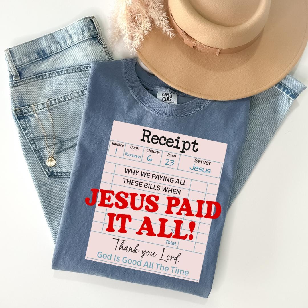 A blue t-shirt with a humorous text printed on it, along with a beige hat and some jeans, all laid out on a white surface.