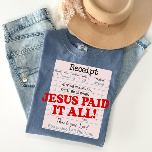 A blue t-shirt with a humorous text printed on it, along with a beige hat and some jeans, all laid out on a white surface.