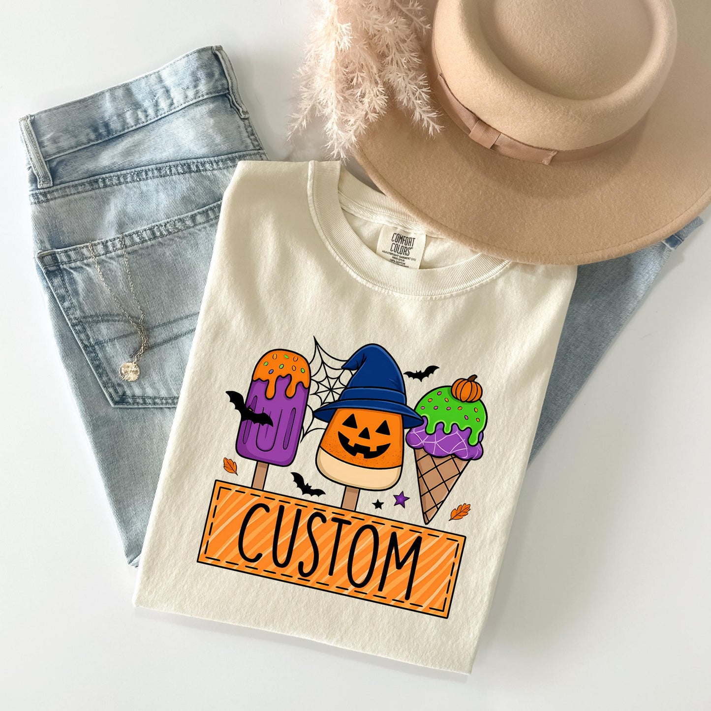 A white t-shirt with a Halloween-themed design, featuring a jack-o-lantern, a witch's hat, and colorful ice cream cones, along with the word "CUSTOM" printed on it. The t-shirt is placed on top of a pair of jeans and a beige hat, creating a casual, festive outfit.