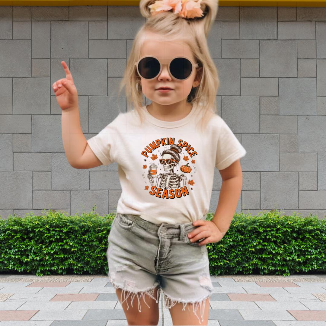 A young girl wearing a white t-shirt with a pumpkin spice design and sunglasses is standing in front of a brick wall.