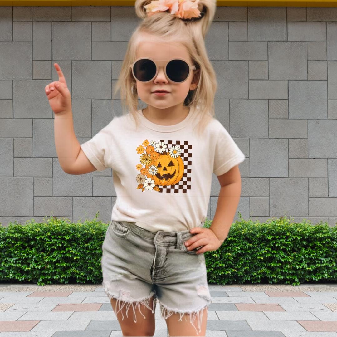 A young girl wearing a white t-shirt with a pumpkin graphic and sunglasses is standing in front of a brick wall.