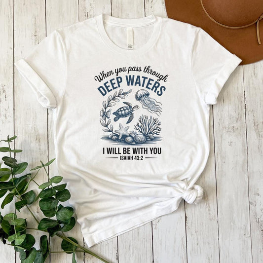A white t-shirt with a graphic design featuring a turtle, jellyfish, and seaweed, along with the text "When you pass through DEEP WATERS, I WILL BE WITH YOU ISAIAH 43:2".