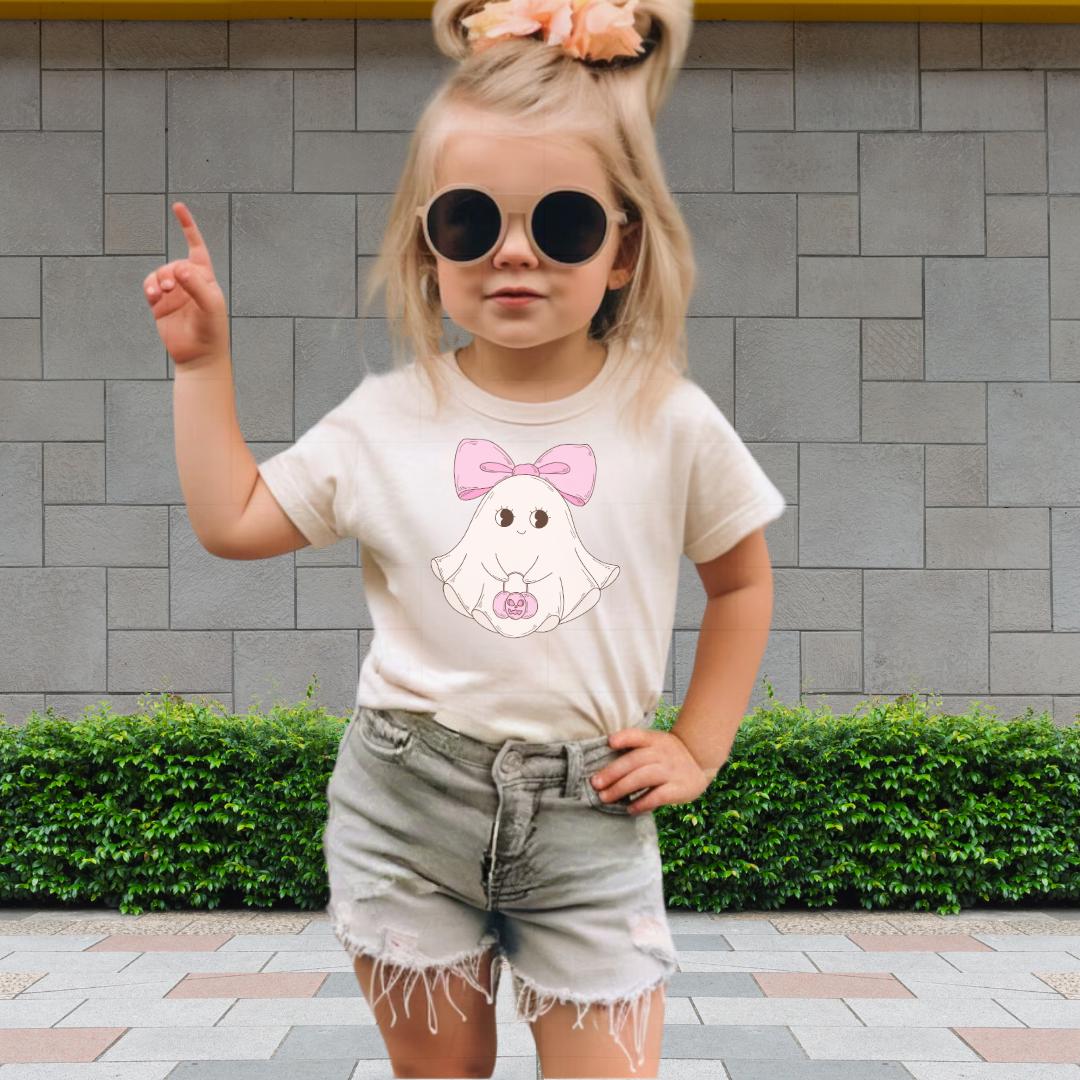 A young girl wearing a white t-shirt with a ghost graphic, sunglasses, and denim shorts is standing in front of a brick wall.