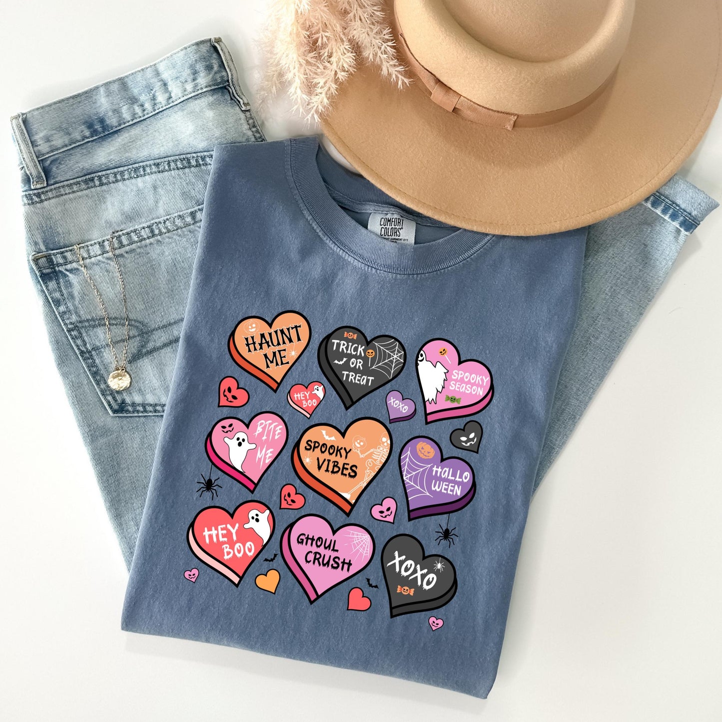 A casual outfit consisting of a blue t-shirt with a graphic design of various Halloween-themed hearts, a pair of jeans, and a beige hat.