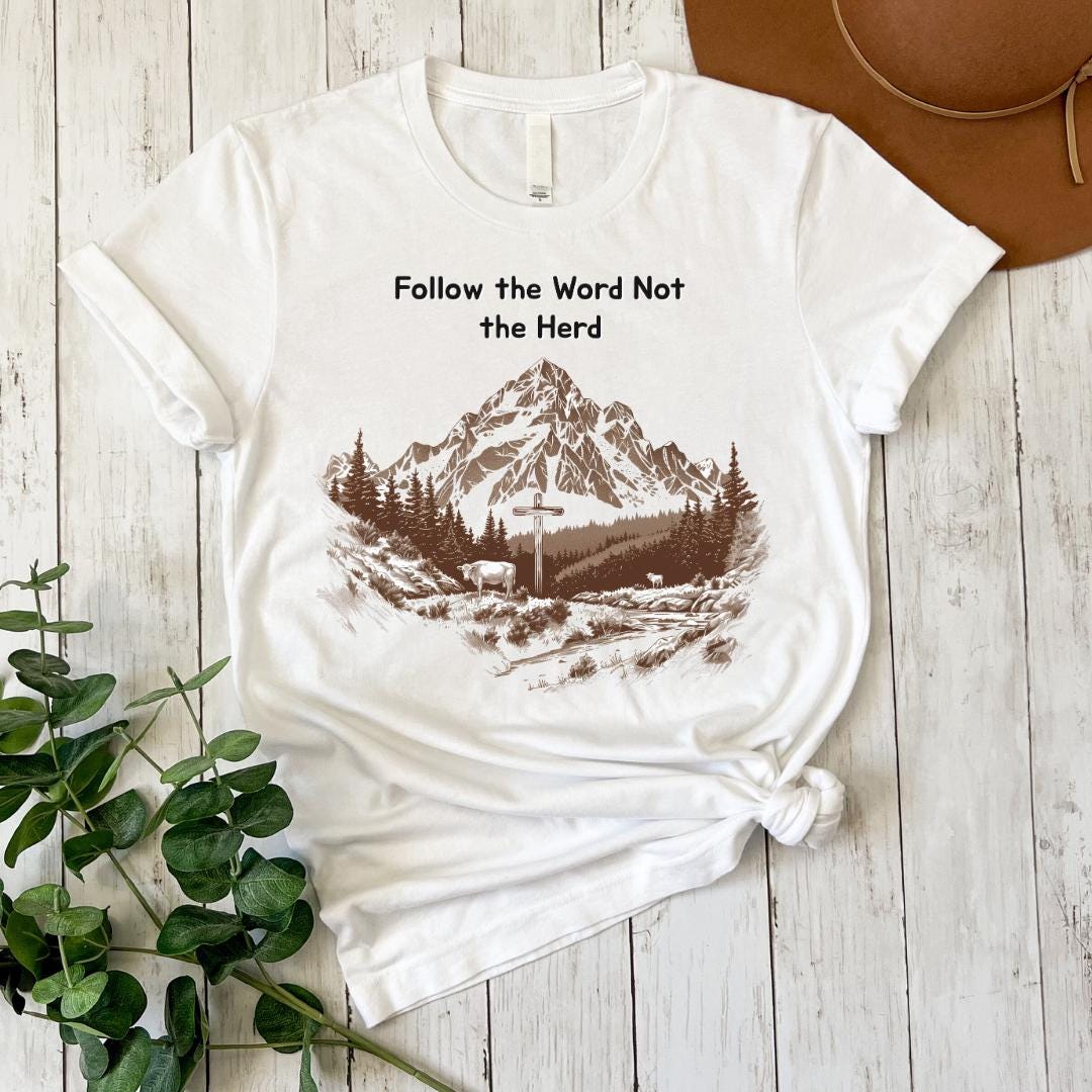 A white t-shirt with a graphic design of a mountain landscape and the text "Follow the Word Not the Herd" printed on it, along with a brown hat placed next to the t-shirt on a wooden surface.