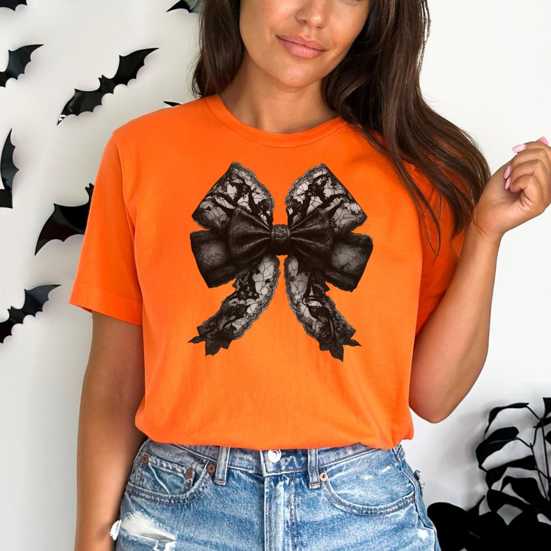 A woman wearing an orange t-shirt with a black bow design on it, standing in front of a wall with black bats hanging on it.