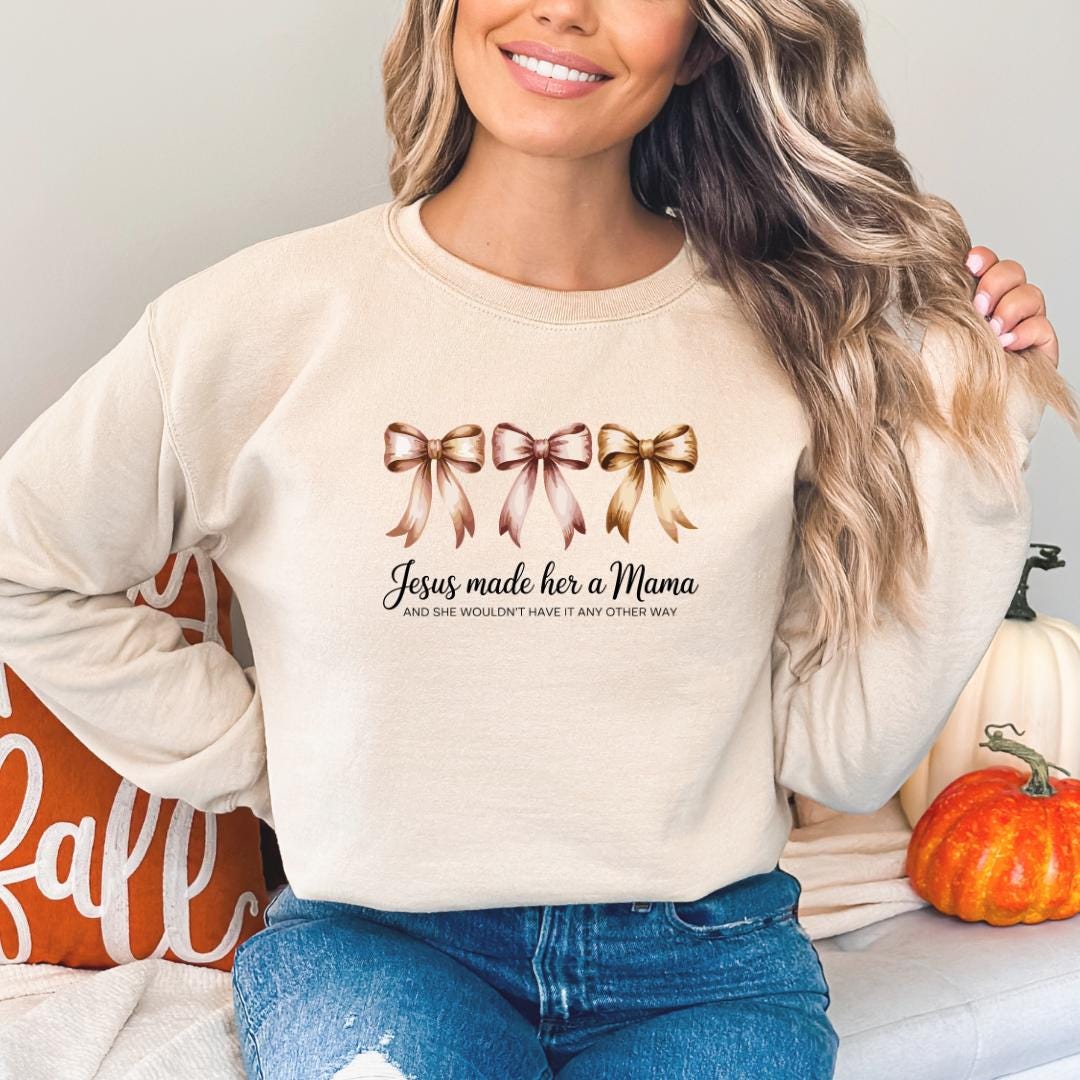 A woman wearing a beige sweater with a graphic design of three bows and text that reads "Jesus made her a Mama and she wouldn't have it any other way".