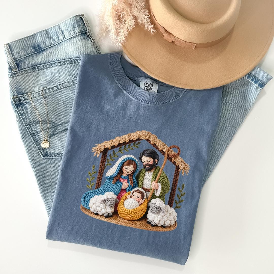 A blue t-shirt with a crochet nativity scene design, a pair of jeans, and a beige hat are arranged on a white background.