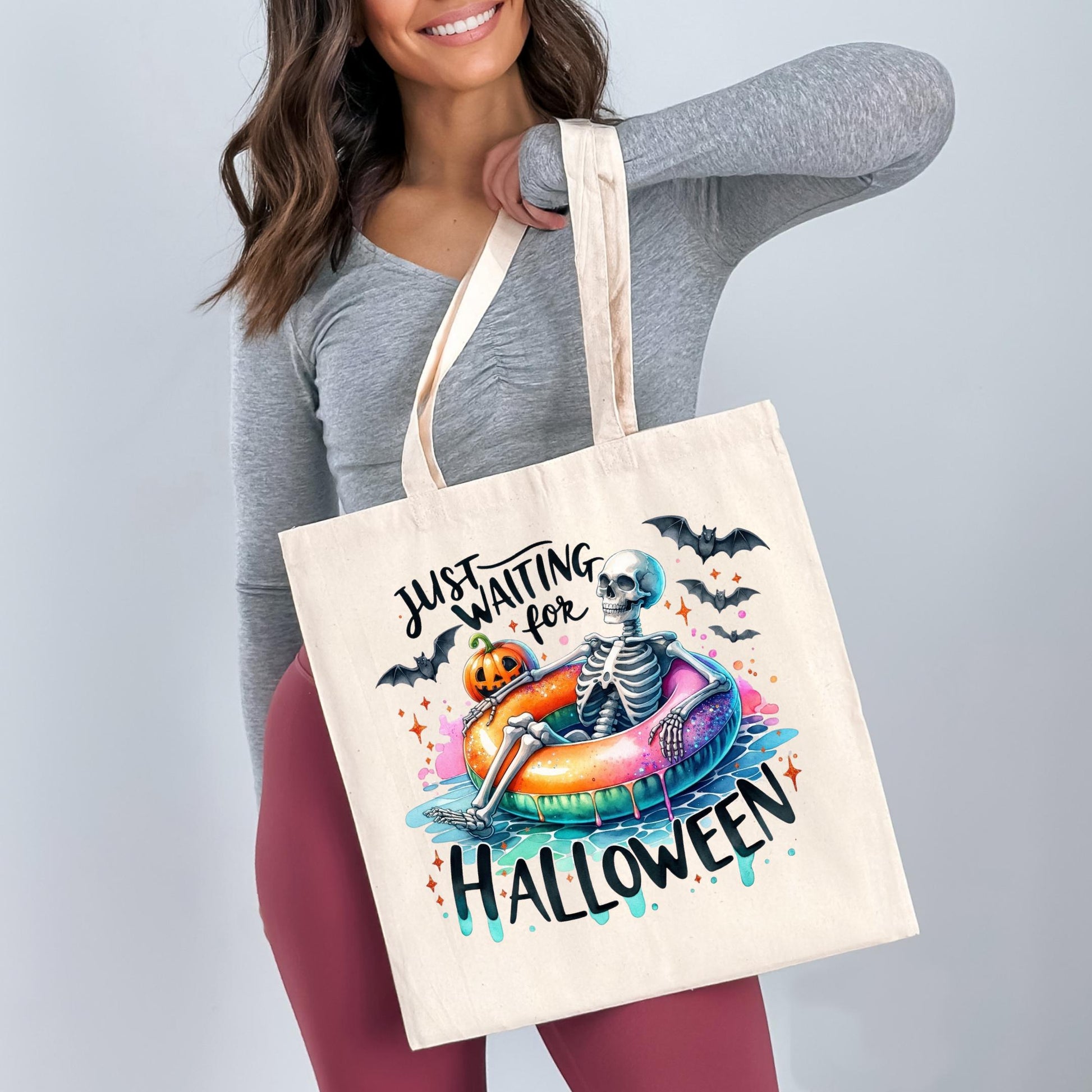 A woman is holding a white tote bag with a Halloween-themed design featuring a skeleton sitting on an inflatable inner tube, surrounded by bats and pumpkins.