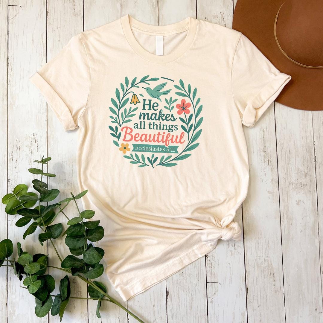 A beige t-shirt with a printed design of a wreath and text, along with a brown hat and some greenery, all placed on a white wooden surface.