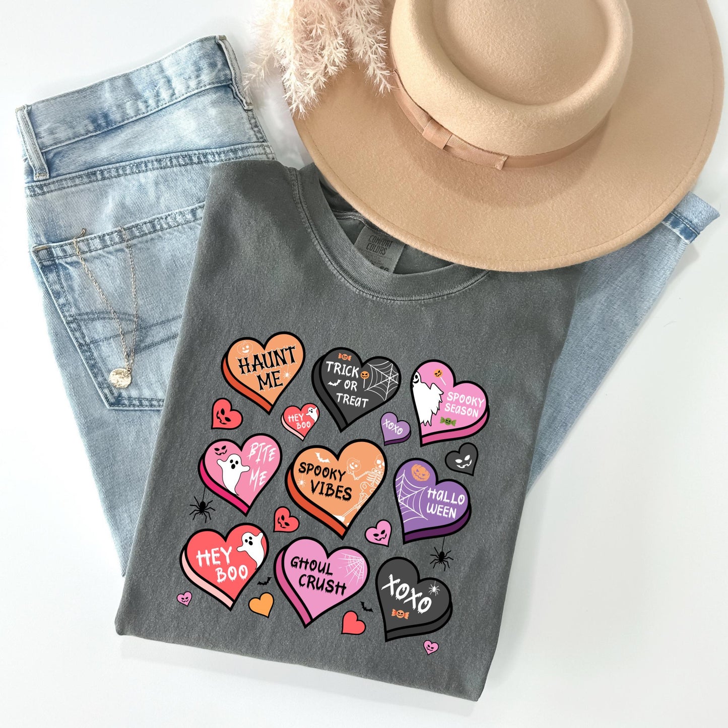 a gray t-shirt with a variety of Halloween-themed heart shapes and text, including phrases like "HAINT ME", "TRICK OR TREAT", "SPOOKY VIBES", "HEY BOO", "CHULO CRUSH", "XOXO", and "HALLO WED". The t-shirt is placed on top of a pair of blue jeans and a beige hat, creating a casual and festive Halloween-inspired outfit.
